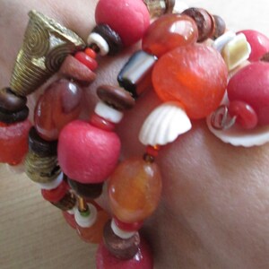 May include: A multi-strand bracelet featuring a mix of red, orange, and brown beads, small white seashell accents, and a conical brass-toned metal charm with etched spiral patterns. The beads vary in shape, size, and texture, including smooth, matte, and translucent finishes strung together on multiple strands.