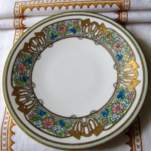 May include: A white porcelain plate with a floral design. The rim features a gold border and a decorative band with pink, blue, and yellow flowers. Gold-colored stylized shapes are interspersed within the floral pattern.