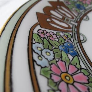 May include: Close-up of a white ceramic plate with a floral design. The plate features a gold rim and a decorative border with pink, blue, and yellow flowers, green leaves, and brown accents. The design is reminiscent of Art Nouveau.