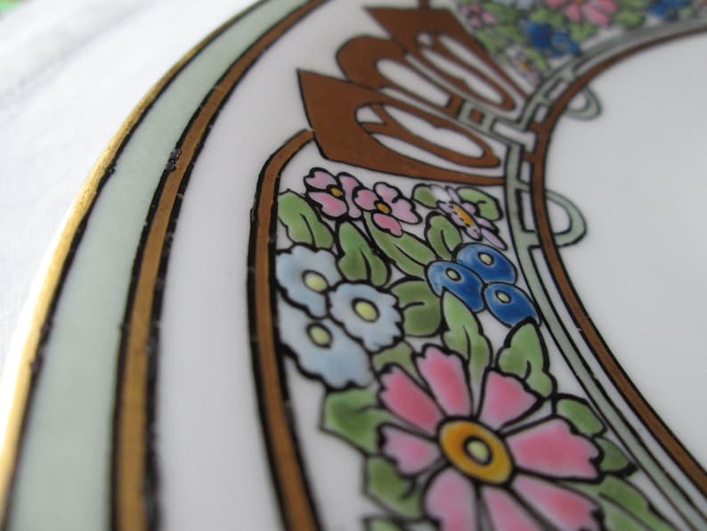 May include: Close-up of a white ceramic plate with a floral design. The plate features a gold rim and a decorative border with pink, blue, and yellow flowers, green leaves, and brown accents. The design is reminiscent of Art Nouveau.