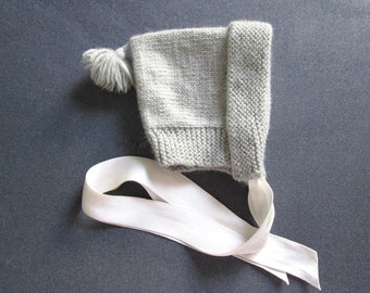 May include: A gray knitted baby bonnet with a white satin ribbon tied around it. The bonnet has a pom-pom on top.