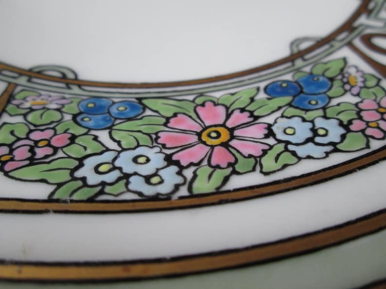 May include: Close-up of a white ceramic plate rim with a floral design. The design features pink, blue, and yellow flowers with green leaves, outlined in black and brown. The plate has a gold-colored border.