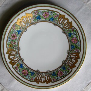 May include: A white ceramic plate with a decorative floral and geometric border. The border features gold accents, pink, blue, and yellow flowers, and a light green outer rim. The plate has a gold trim.