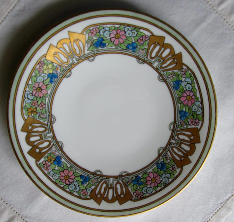 May include: A white ceramic plate with a decorative floral and geometric border. The border features gold accents, pink, blue, and yellow flowers, and a light green outer rim. The plate has a gold trim.