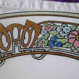 May include: Close-up of a white plate with an Art Nouveau design. The plate features gold-colored lettering and a floral border with pink, blue, and yellow flowers. The design is outlined in black and green.