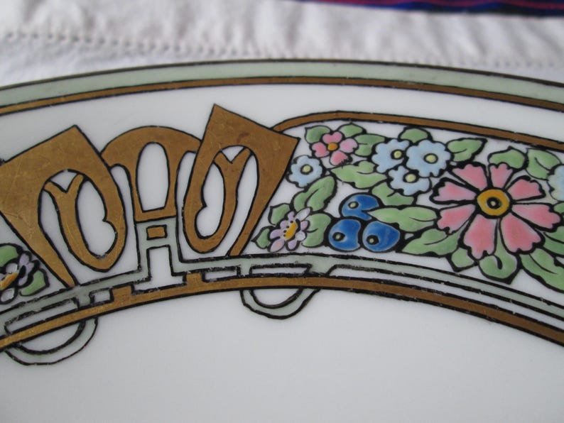 May include: Close-up of a white plate with an Art Nouveau design. The plate features gold-colored lettering and a floral border with pink, blue, and yellow flowers. The design is outlined in black and green.
