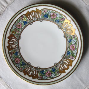 May include: White ceramic plate with a floral design. The plate features a gold rim and gold accents around the edge. The floral pattern includes pink, blue, and yellow flowers.