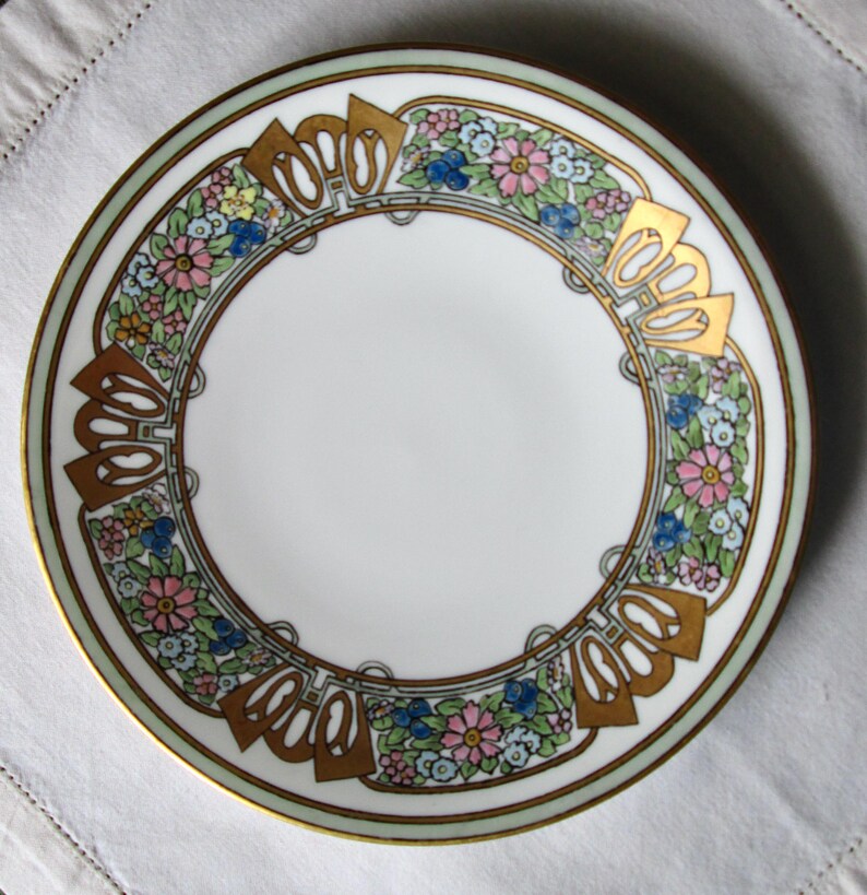 May include: White ceramic plate with a floral design. The plate features a gold rim and gold accents around the edge. The floral pattern includes pink, blue, and yellow flowers.