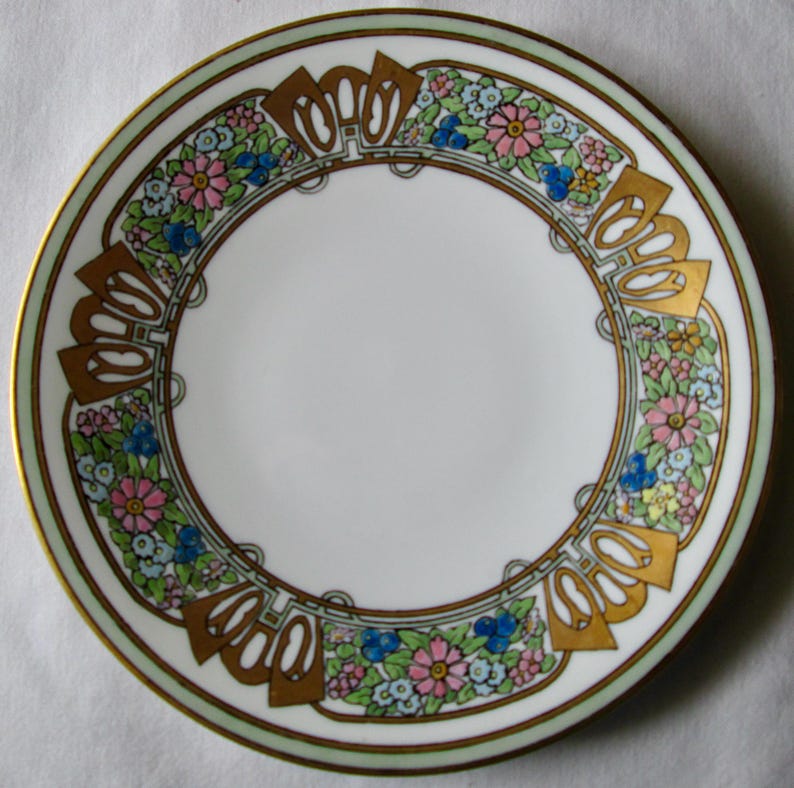 May include: A white porcelain plate with a decorative floral border. The border features pink, blue, and yellow flowers with gold accents and a light green rim. The plate has a gold trim.