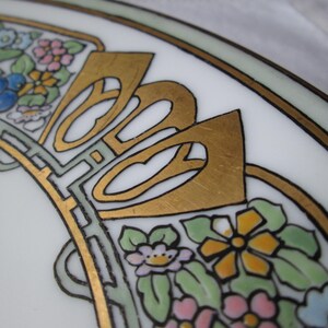 May include: Close-up of a white ceramic plate with an Art Nouveau design. The plate features gold accents, black outlines, and colorful floral patterns in pink, blue, yellow, and green. The design is symmetrical and ornate.