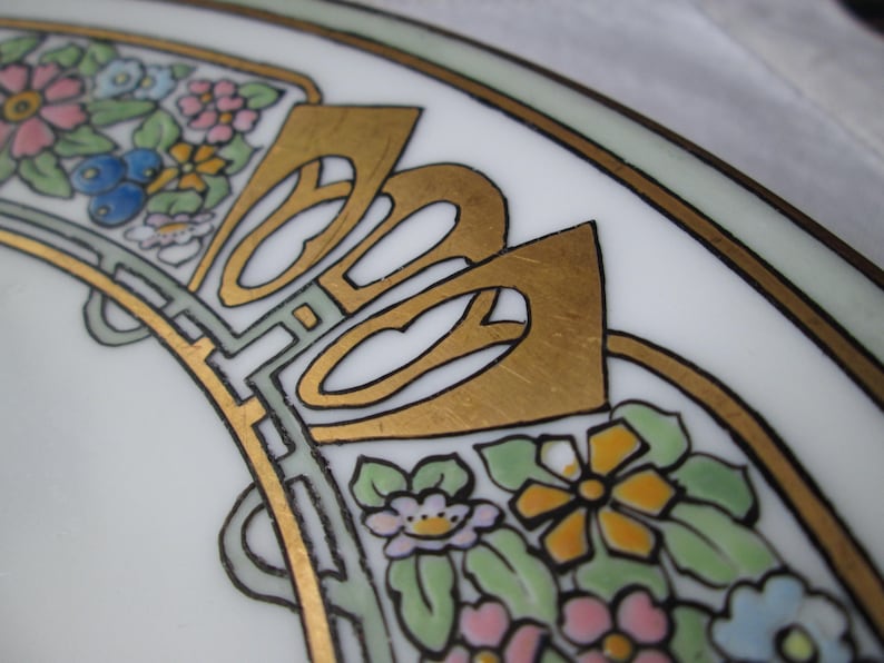 May include: Close-up of a white ceramic plate with an Art Nouveau design. The plate features gold accents, black outlines, and colorful floral patterns in pink, blue, yellow, and green. The design is symmetrical and ornate.