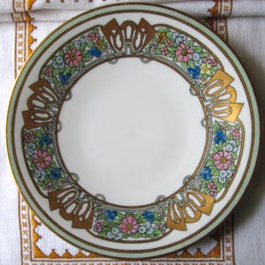May include: A white ceramic plate with a floral design. The plate features a border of pink, blue, and yellow flowers, with gold and green accents. The plate has a gold rim and a decorative pattern.