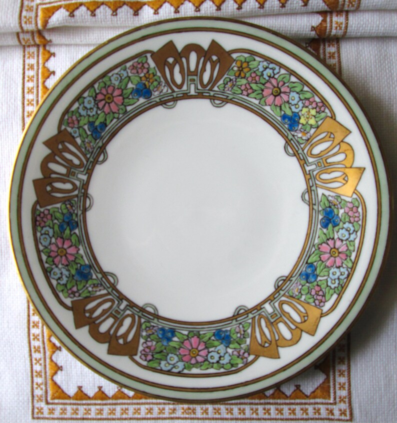 May include: A white ceramic plate with a floral design. The plate features a border of pink, blue, and yellow flowers, with gold and green accents. The plate has a gold rim and a decorative pattern.