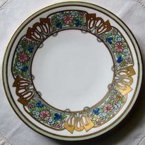 May include: A white porcelain plate with a decorative floral border. The border features pink, blue, and yellow flowers, gold accents, and a light green edge. The plate has a gold rim.
