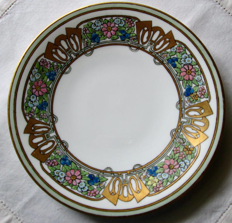 May include: A white porcelain plate with a decorative floral border. The border features pink, blue, and yellow flowers, gold accents, and a light green edge. The plate has a gold rim.