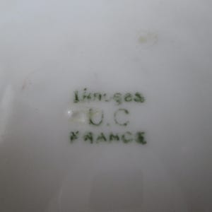 May include: Close-up of a white porcelain surface with green text that reads "Limoges U.C. FRANCE". The text is slightly blurred. The porcelain has a glossy finish.