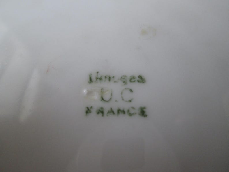 May include: Close-up of a white porcelain surface with green text that reads "Limoges U.C. FRANCE". The text is slightly blurred. The porcelain has a glossy finish.