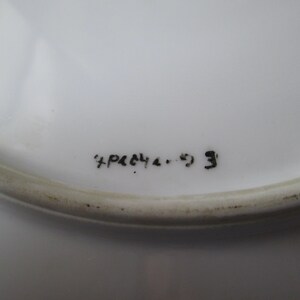 May include: Close-up of a white ceramic plate with a slightly raised edge. The plate has black handwritten text that reads "+P&C4 4. 9 3". The plate is clean and appears to be in good condition.