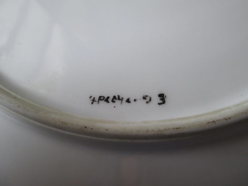 May include: Close-up of a white ceramic plate with a slightly raised edge. The plate has black handwritten text that reads "+P&C4 4. 9 3". The plate is clean and appears to be in good condition.