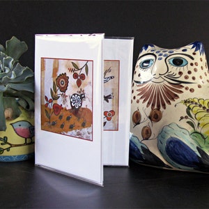 May include: Two greeting cards with colorful, whimsical illustrations of birds and flowers. The cards are displayed on a black surface next to a ceramic cat figurine with a colorful floral design.