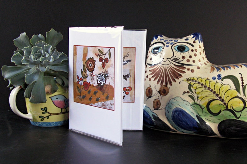 May include: Two greeting cards with colorful, whimsical illustrations of birds and flowers. The cards are displayed on a black surface next to a ceramic cat figurine with a colorful floral design.