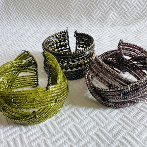 May include: Three beaded bracelets in shades of green, bronze, and burgundy. The bracelets are made of small beads strung together on wire, creating a textured and layered effect. The bracelets are displayed on a white, textured surface.