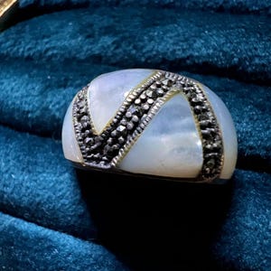 Può includere: Un anello perlato con una fascia argentata e un design decorativo in pietra nera. L'anello ha una forma arrotondata ed è posto su uno sfondo verde acqua scuro. Il design dell'anello presenta un motivo a forma di V.