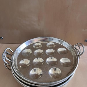 Puede incluir: Una pila de tres platos plateados para servir caracoles. Cada plato tiene 12 huecos empotrados y dos asas en forma de lazo. Los platos son redondos y parecen ser de metal.