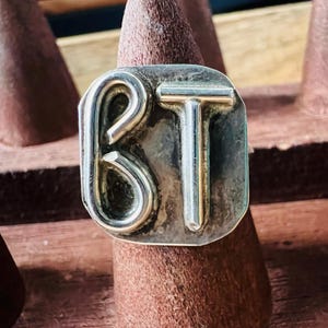 May include: A silver-toned ring with the letters "BT" in a bold, stylized font. The ring has a rectangular face with rounded corners. The letters are raised against a slightly textured background. The ring is displayed on a brown, cone-shaped stand.