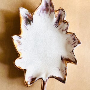 以下が含まれることがあります： 白い内側とひび割れのある釉薬が施された、葉の形をしたセラミック皿。縁は金でトリミングされ、先端は茶色と紫色のメタリック仕上げです。この皿は葉のようにデザインされています。