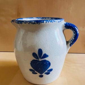 Könnte beinhalten: Ein weißer Keramikkrug mit blauem Herzmuster und blauem Griff. Der Rand und der Griff sind blau eingefasst. Der Krug hat eine Craquelé-Glasur und steht vor einem hellbraunen Hintergrund.