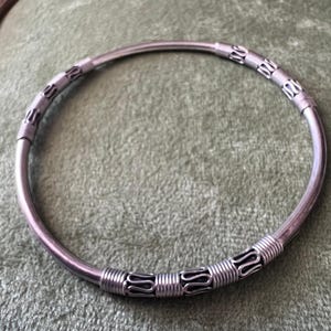 Puede incluir: Un brazalete de color plateado. El brazalete tiene un diseño circular simple con secciones decorativas con intrincados envoltorios de alambre y patrones estilizados. El brazalete se exhibe sobre una superficie verde.
