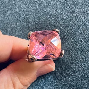 May include: A large, square-cut pink gemstone ring. The ring features a faceted, light pink stone set in a silver-toned metal band. The gemstone is held in place by four prongs. The ring is held by a hand.