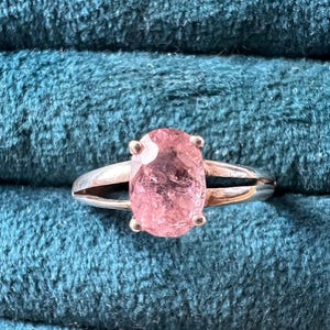 May include: A silver ring featuring a large, oval-cut pink gemstone. The gemstone is set with four prongs and has a split shank design. The ring is displayed against a teal, textured background.