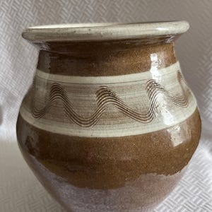 Peut inclure: Un vase en céramique fait à la main avec un corps arrondi et une large ouverture. Le vase présente une base marron, une bande crème avec des motifs ondulés et un bord crème. La poterie a un aspect rustique et texturé.