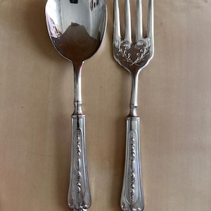 Puede incluir: Un juego de cuchara y tenedor de plata. La cuchara tiene un cuenco grande y ovalado, y el tenedor tiene cuatro púas. Ambos utensilios tienen mangos ornamentados y detallados con un patrón decorativo. El juego se muestra sobre una superficie marrón claro.