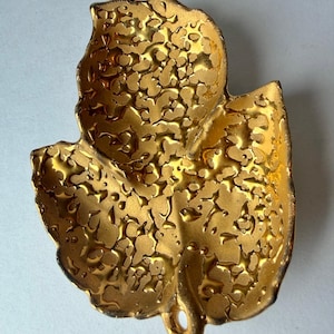 May include: A gold-colored decorative dish shaped like a leaf with a textured, uneven surface. The metallic finish has a reflective quality with raised, organic patterns across the interior, and a small circular hole at the base of the stem.