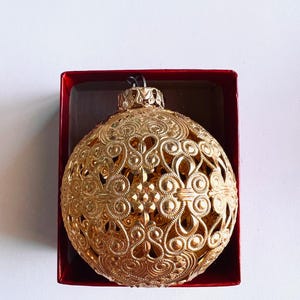 Op de afbeelding: Een goudkleurige, sierlijke kerstbal met een gedetailleerd, opengewerkt ontwerp. De bolvormige ornament bevindt zich in een rode doos, wat een feestelijke presentatie creëert. De ingewikkelde patronen en metallic afwerking suggereren een vintage of handgemaakte stijl.