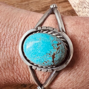 May include: A silver cuff bracelet featuring a large, oval turquoise stone. The turquoise is a vibrant blue-green with brown matrix patterns. The bracelet has a geometric design with decorative elements.