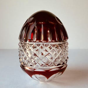 May include: A clear glass jar with a ruby red lid and base. The jar's body features a diamond-cut pattern, while the lid has a rounded, textured design. The jar is likely used for storing small items.