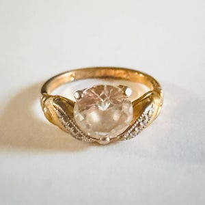 Può includere: Un anello dorato con una grande gemma rotonda e trasparente come elemento centrale. La fascia presenta dettagli decorativi e la gemma è tenuta in posizione da quattro griffe. Piccole pietre trasparenti accentuano i lati dell'anello.