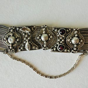 Può includere: Un braccialetto argentato con maglie geometriche ornate e decorazioni centrali. Il braccialetto ha un design a catena con più fili ed elementi decorativi, tra cui quelli che sembrano essere pietre preziose.