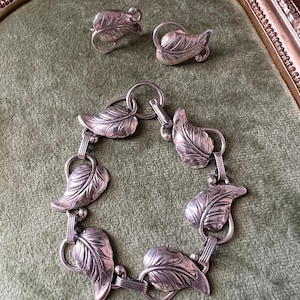 Puede incluir: Un conjunto de joyas plateadas que incluye una pulsera y pendientes a juego. La pulsera está compuesta por eslabones en forma de hoja, y los pendientes tienen un diseño similar. El conjunto se exhibe sobre una superficie de terciopelo verde.