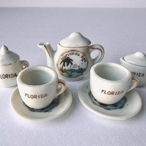 May include: A miniature ceramic tea set featuring a teapot, two cups with saucers, a sugar bowl, and a creamer. Each white piece is decorated with brown text reading FLORIDA and a small graphic of a palm tree and a sailboat on the teapot.