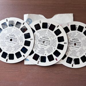 May include: Three vintage View-Master reels featuring "Dennis the Menace" stories. Each circular reel has a series of small windows with images and text. The reels are white with black text and are arranged on a wooden surface.