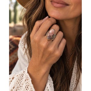 May include: An oval-shaped ring featuring a multi-colored stone with swirling patterns of pink, white, and grey. The stone is set in a metallic band with a textured, hammered finish and a thin gold-toned border surrounding the central gemstone.