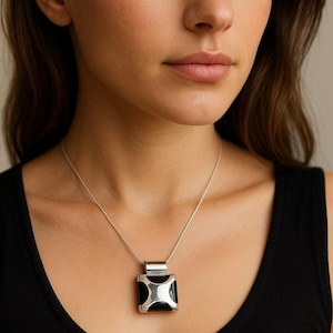 Puede incluir: Collar con colgante cuadrado plateado con diseño geométrico negro y plateado. El colgante cuelga de una delicada cadena plateada. La mujer de la imagen lleva una camiseta sin mangas negra.