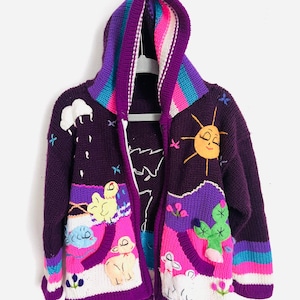 May include: A purple hooded sweater with colorful landscape and animal appliqués. The sweater features a smiling sun, clouds, a cactus, and sheep. The hood has rainbow stripes, and the sweater is hanging on a white hanger.