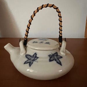 May include: White ceramic teapot with a woven handle and a lid. The teapot is decorated with blue leaf designs and Japanese characters. The handle is made of woven brown and black material.