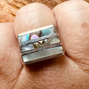 May include: A silver ring with four rectangular sections. Each section features a different iridescent abalone shell inlay, displaying shades of blue, green, and pink. The ring is worn on a finger.